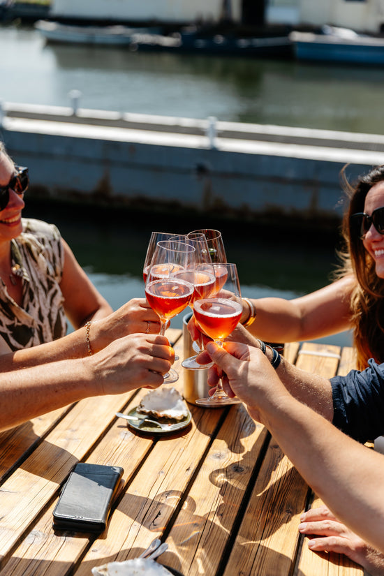 People toasting with glasses of pink rose Yvonne Belhomme Champagne on a wooden dock by a body of water.