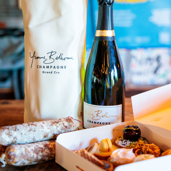 A champagne bottle and a branded canvas tote bag Yvonne Belhomme sit on a wooden table beside an open gift box with pastries, with a blurred indoor background.