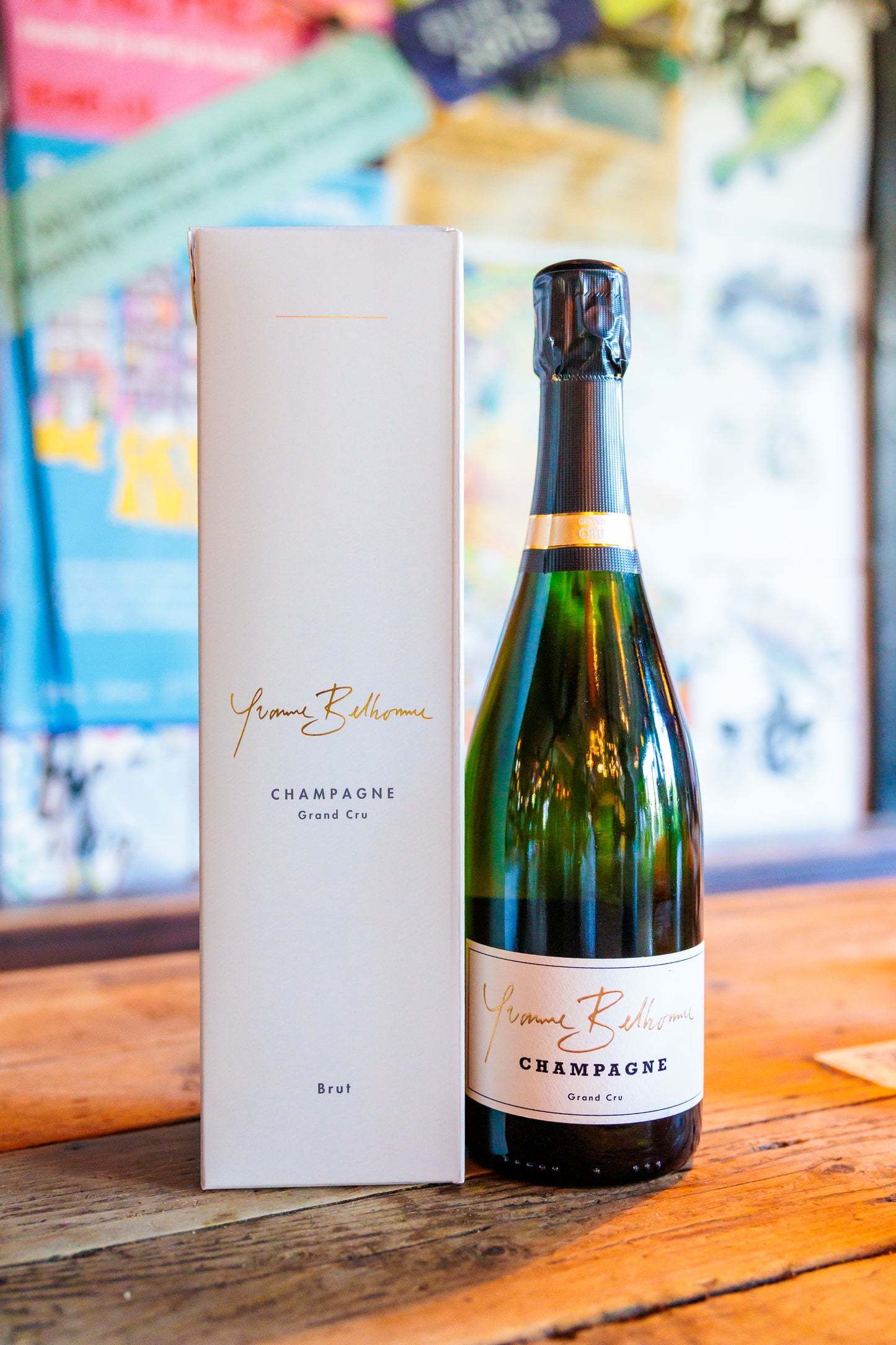 A bottle of Yvonne Belhomme champagne next to its tall white presentation box, set on a rustic wooden table with a softly blurred background.