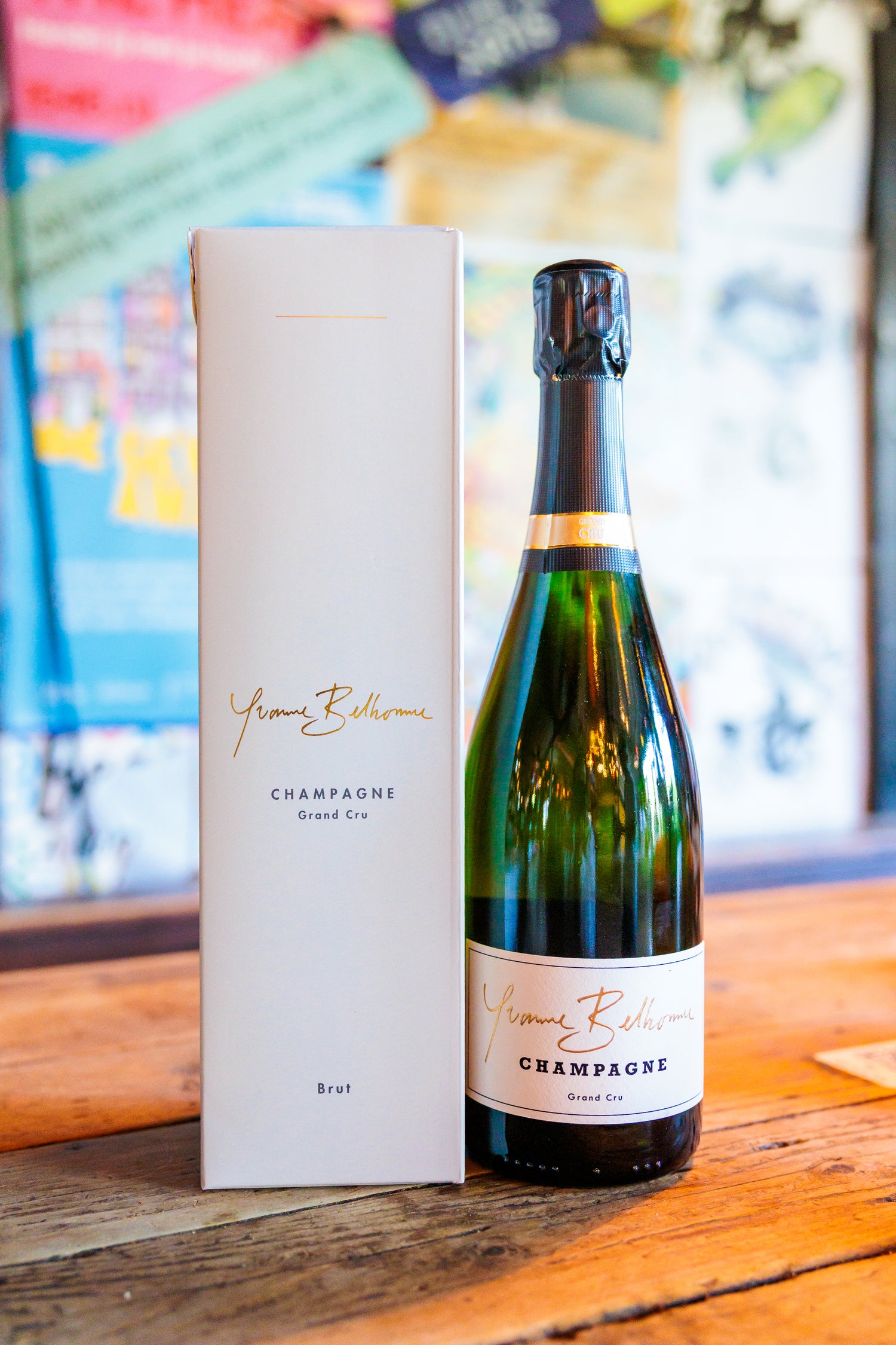 A bottle of Yvonne Belhomme champagne next to its tall white presentation box, set on a rustic wooden table with a softly blurred background.