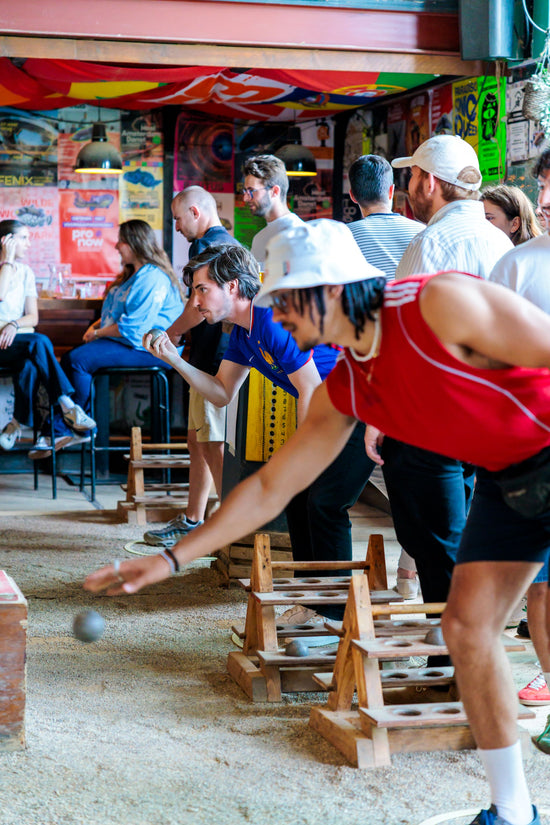 A man in a red sleeveless top and white cap throws a petanque in an indoor game, with several other people and colorful wall murals in the background.