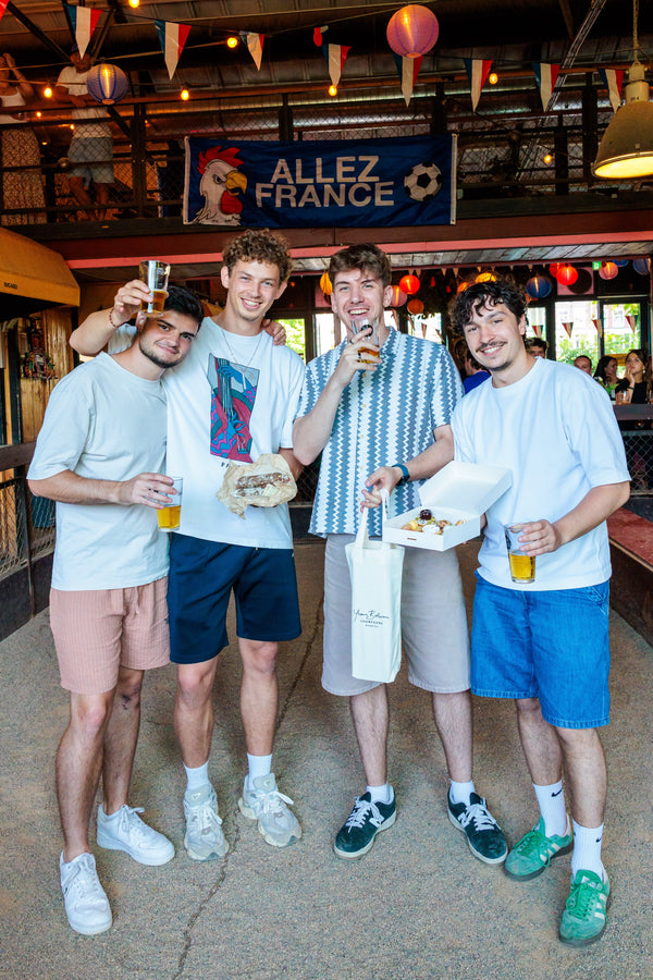 Four young men standing together in a festive indoor venue with French decorations, holding drinks and food, with a banner reading 'ALLEZ FRANCE' in the background.