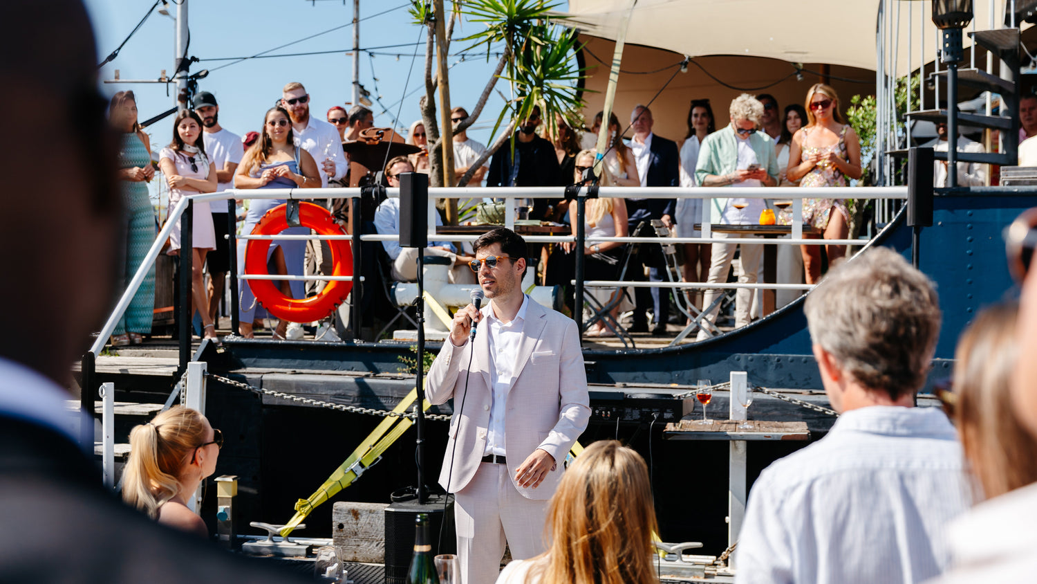 Man founder of Yvonne Belhomme Champagne in a pink suit speaking on a boat with people watching